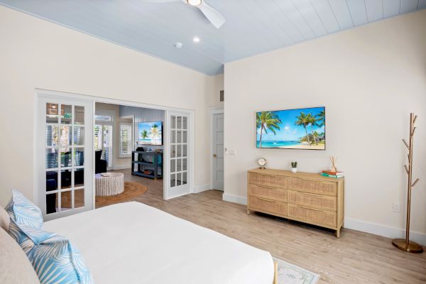 A bedroom with a bed, dresser, TV displaying a beach scene, and adjacent living area with glass doors and additional TV.