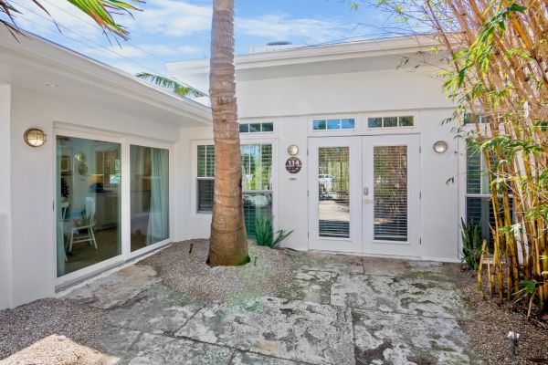 The image shows a modern patio with stone flooring, a palm tree, and a white building with large windows and glass doors.