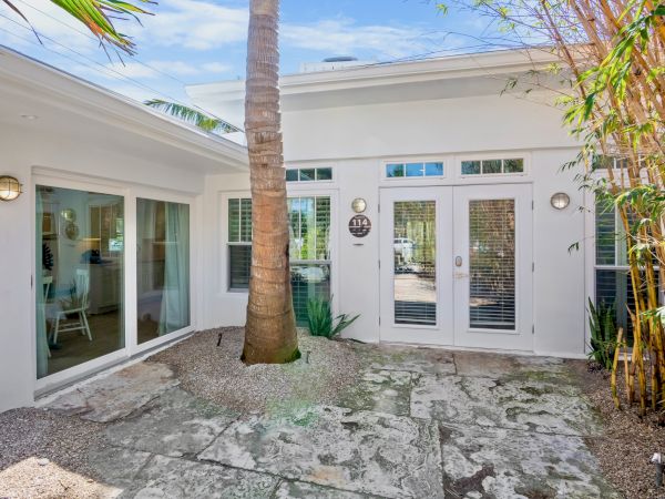 The image shows a modern patio with stone flooring, a palm tree, and a white building with large windows and glass doors.