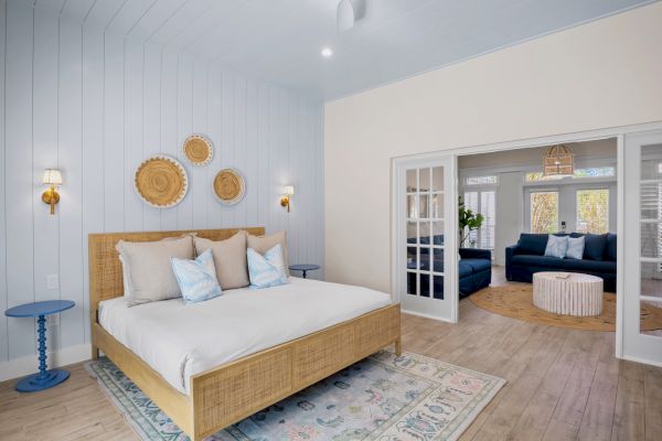 A cozy bedroom with a wooden bed, patterned rug, decorative wall pieces, and a living area with a blue sofa visible through double doors.