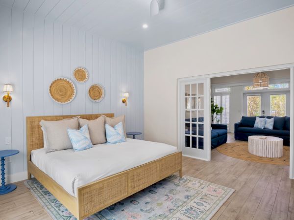 A cozy bedroom with a wooden bed, patterned rug, decorative wall pieces, and a living area with a blue sofa visible through double doors.