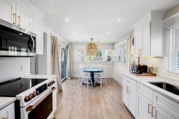 A modern kitchen with white cabinets, wooden floor, and a dining area. There's a round table with chairs by a window at the back.