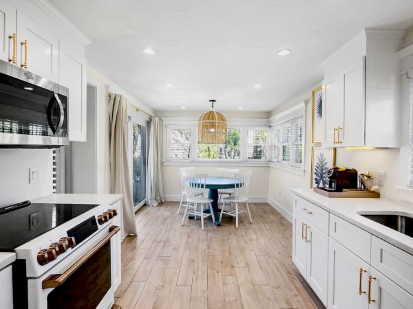 A modern kitchen with white cabinets, wooden floor, and a dining area. There's a round table with chairs by a window at the back.