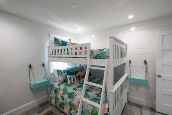 A cozy bedroom with a white bunk bed, tropical-themed bedding, floating shelves, and a ladder, set in a well-lit, minimalistic space.