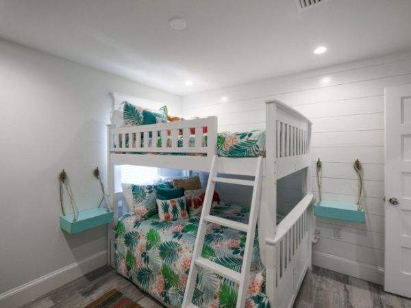 A cozy bedroom with a white bunk bed, tropical-themed bedding, floating shelves, and a ladder, set in a well-lit, minimalistic space.