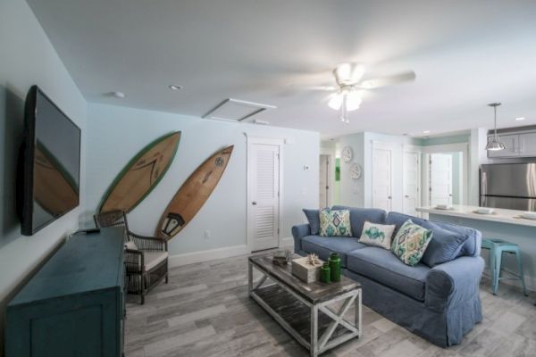 A cozy living room with a blue sofa, surfboards on the wall, TV, and a kitchen area with stools and stainless steel appliances.