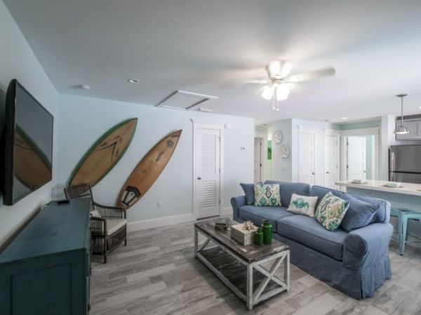 A cozy living room with a blue sofa, surfboards on the wall, TV, and a kitchen area with stools and stainless steel appliances.