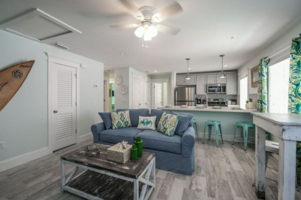 A cozy living area with a blue couch, patterned cushions, coffee table, open kitchen, and surfboard on the wall, with bright decor accents.