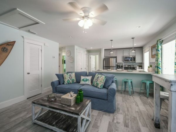 A cozy living area with a blue couch, patterned cushions, coffee table, open kitchen, and surfboard on the wall, with bright decor accents.