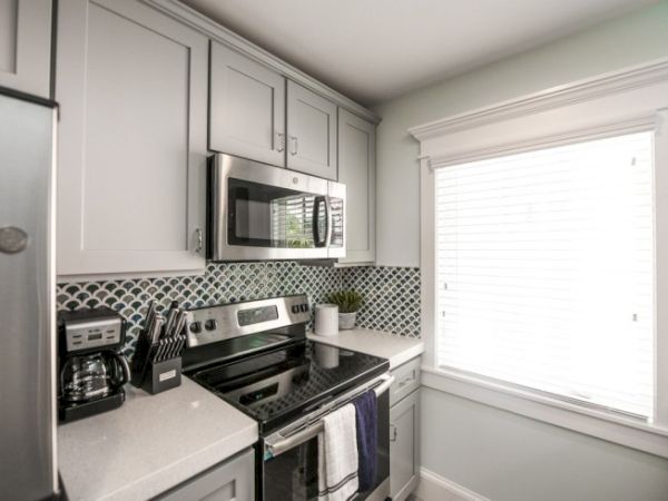 A modern kitchen with white cabinets, stainless steel appliances, a fish-scale backsplash, and a window with blinds.