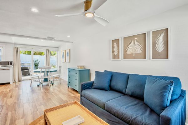 A modern living room with a blue sofa, palm wall art, wooden flooring, and a small dining area near the windows.