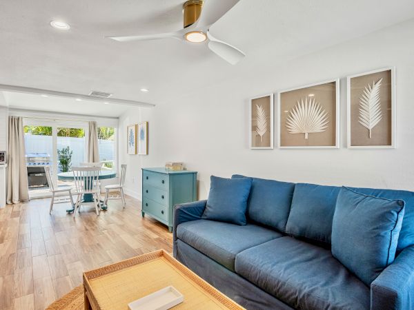 A modern living room with a blue sofa, palm wall art, wooden flooring, and a small dining area near the windows.