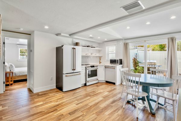 A modern kitchen with wooden flooring, a fridge, stove, table, and chairs, adjacent to a bedroom and a patio with a grill.