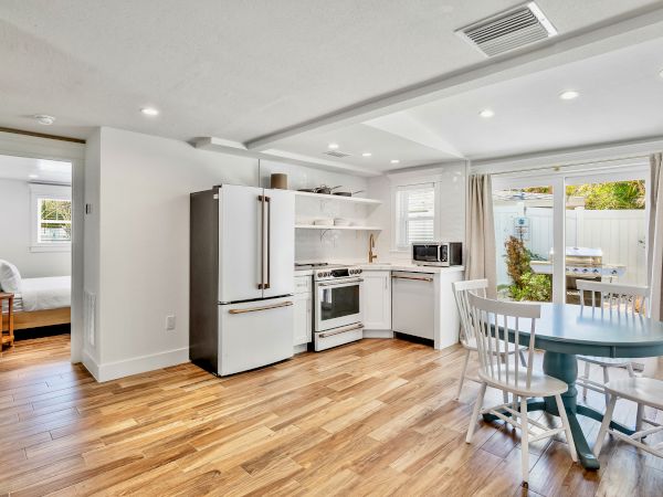 A modern kitchen with wooden flooring, a fridge, stove, table, and chairs, adjacent to a bedroom and a patio with a grill.