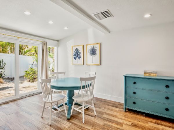 A bright room with a round table, three chairs, a blue dresser, and two framed plants. Glass doors open to a garden area.