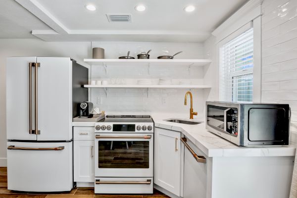 A modern kitchen features a fridge, stove, microwave, coffee maker, white cabinets, and shelves with cookware and dishes.