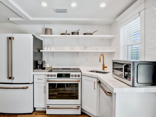 A modern kitchen features a fridge, stove, microwave, coffee maker, white cabinets, and shelves with cookware and dishes.