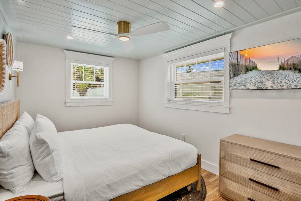 A small bedroom features a bed with white linens, two windows, a ceiling fan, a dresser, and a beach-themed artwork.