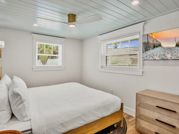 A small bedroom features a bed with white linens, two windows, a ceiling fan, a dresser, and a beach-themed artwork.