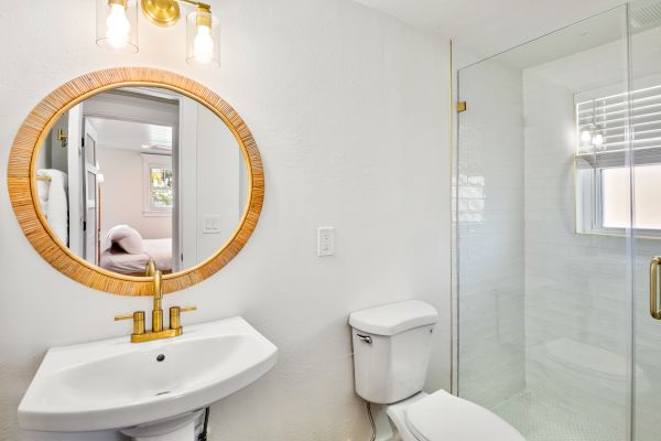 A modern bathroom with a round mirror, sink, toilet, and glass shower enclosure featuring a gold faucet and light fixture.
