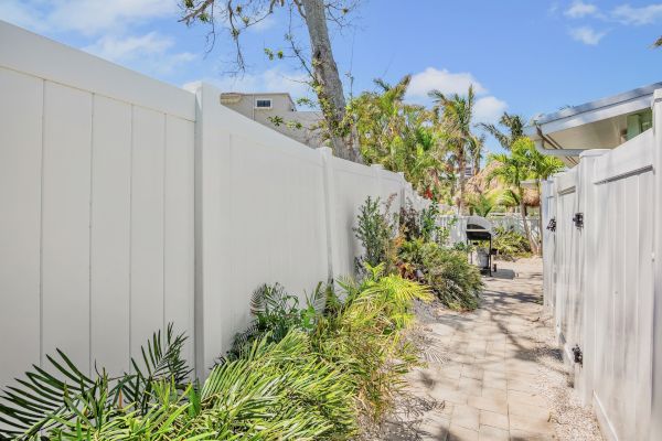A narrow paved path lined with plants, white fences on both sides, and a grill in the distance under a blue sky.