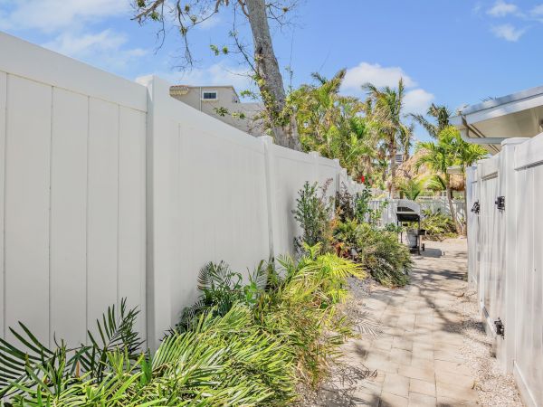 A narrow paved path lined with plants, white fences on both sides, and a grill in the distance under a blue sky.