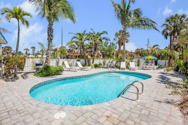 A sunny outdoor swimming pool area with lounge chairs, palm trees, and a table with an umbrella, creating a tropical, relaxing setting.