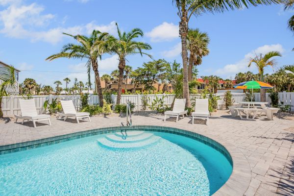 A swimming pool with lounge chairs, palm trees, and a table with an umbrella in a sunny outdoor setting.