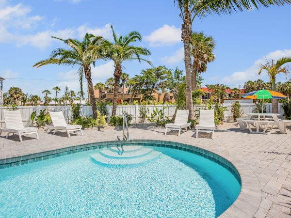 A swimming pool with lounge chairs, palm trees, and a table with an umbrella in a sunny outdoor setting.