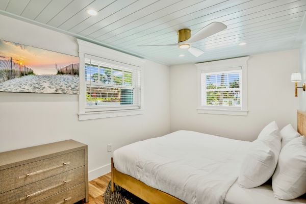 A cozy bedroom with a double bed, ceiling fan, dresser, windows, and beach-themed artwork on the wall.