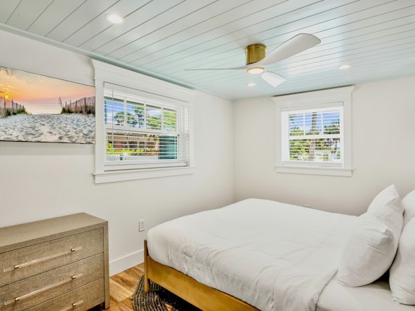 A cozy bedroom with a double bed, ceiling fan, dresser, windows, and beach-themed artwork on the wall.