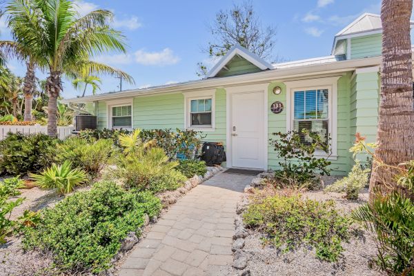 A small, green cottage with white trim is surrounded by palm trees and lush landscaping, under a bright blue sky.