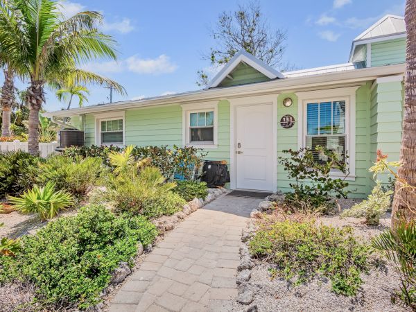 A small, green cottage with white trim is surrounded by palm trees and lush landscaping, under a bright blue sky.