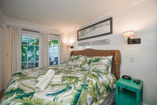 A cozy bedroom with a tropical theme, featuring a bed with palm leaf bedding, coastal decor, and natural light through sliding doors.