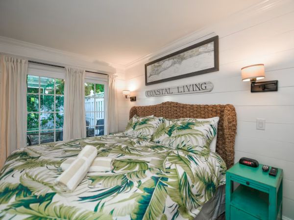 A cozy bedroom with a tropical theme, featuring a bed with palm leaf bedding, coastal decor, and natural light through sliding doors.