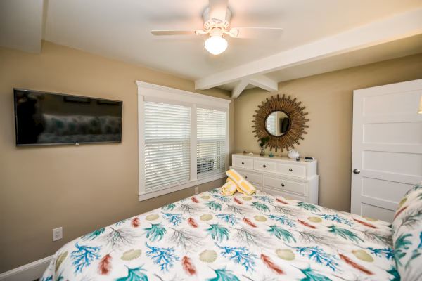 A cozy bedroom with a colorful patterned bedspread, a dresser with a mirror, a ceiling fan, and a wall-mounted TV.