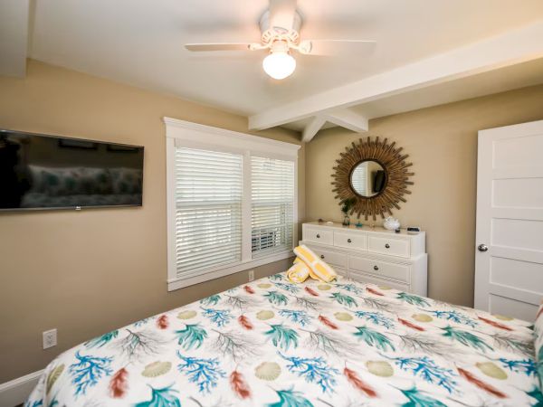 A cozy bedroom with a colorful patterned bedspread, a dresser with a mirror, a ceiling fan, and a wall-mounted TV.