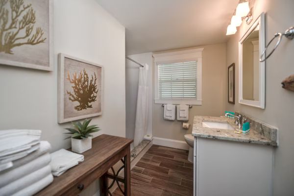 A bathroom with a shower, sink, and towels. Decor includes plant and art on walls. Natural light from a window with blinds.