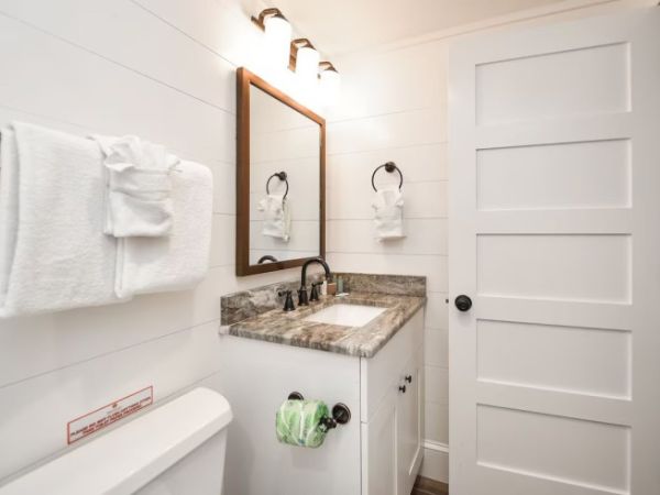 A modern bathroom with a marble countertop, sink, mirror, and white door. Towels hang on the wall and toilet paper rolls are visible.