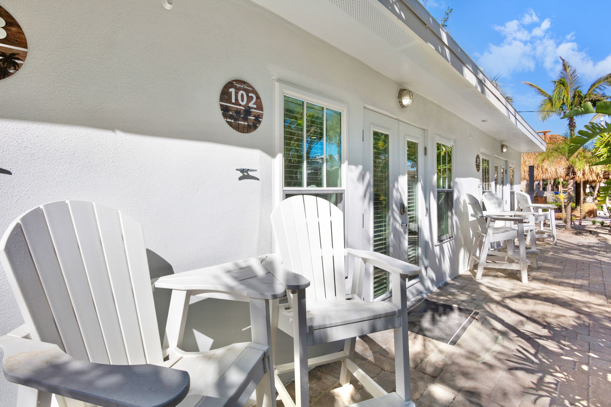 A sunny patio area with white Adirondack chairs outside a building with room numbers on the wall. Palm trees and a clear sky in the background.