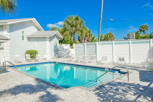 A clear swimming pool surrounded by lounge chairs and a white fence, with palm trees and a sunny blue sky in the background.
