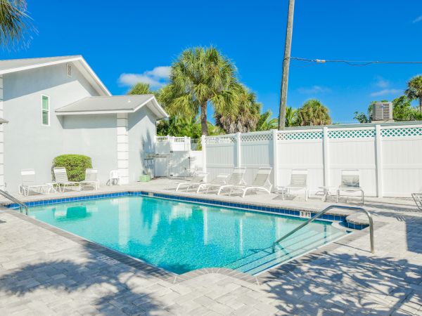 A clear swimming pool surrounded by lounge chairs and a white fence, with palm trees and a sunny blue sky in the background.
