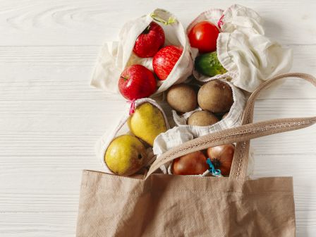 Reusable cloth bags filled with apples, a tomato, a cucumber, potatoes, pears, and onions, inside a brown tote bag on a white surface.