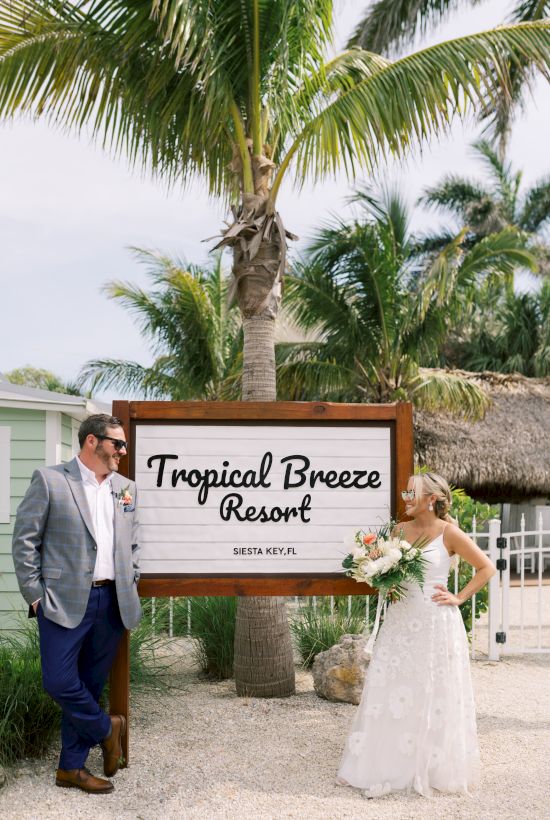 A couple stands by a "Tropical Breeze Resort" sign in Siesta Key, FL, with palm trees in the background, both dressed for a wedding.