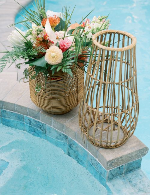 A wicker basket with lush flowers and greenery sits beside a pool, alongside a decorative wooden lantern.