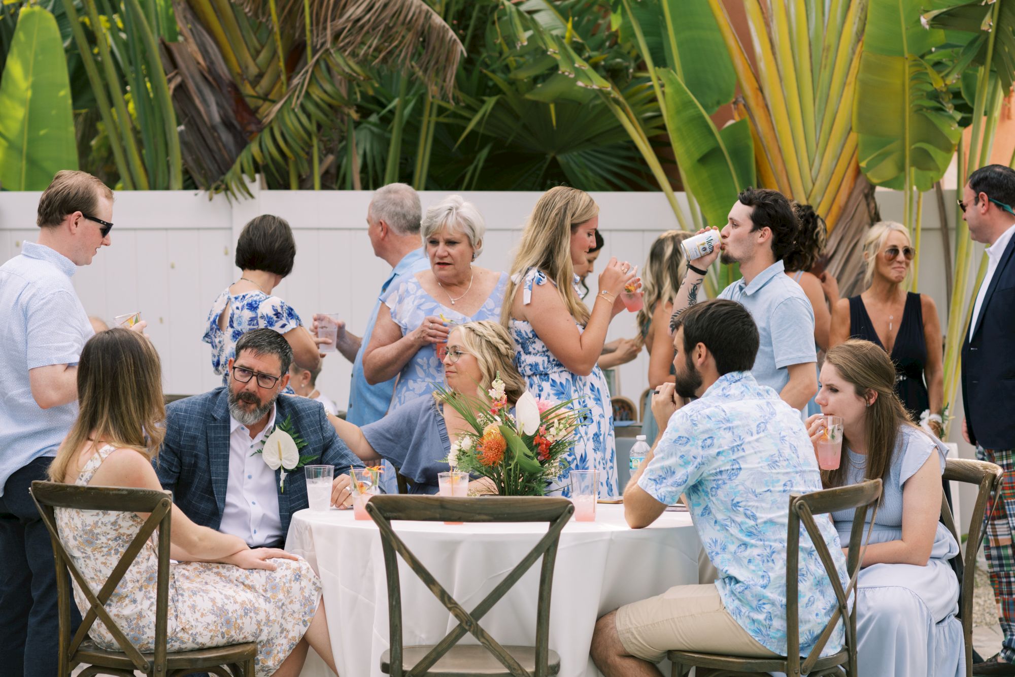 A group of people are gathered at an outdoor event, chatting and enjoying drinks at a table surrounded by lush greenery, appearing relaxed.