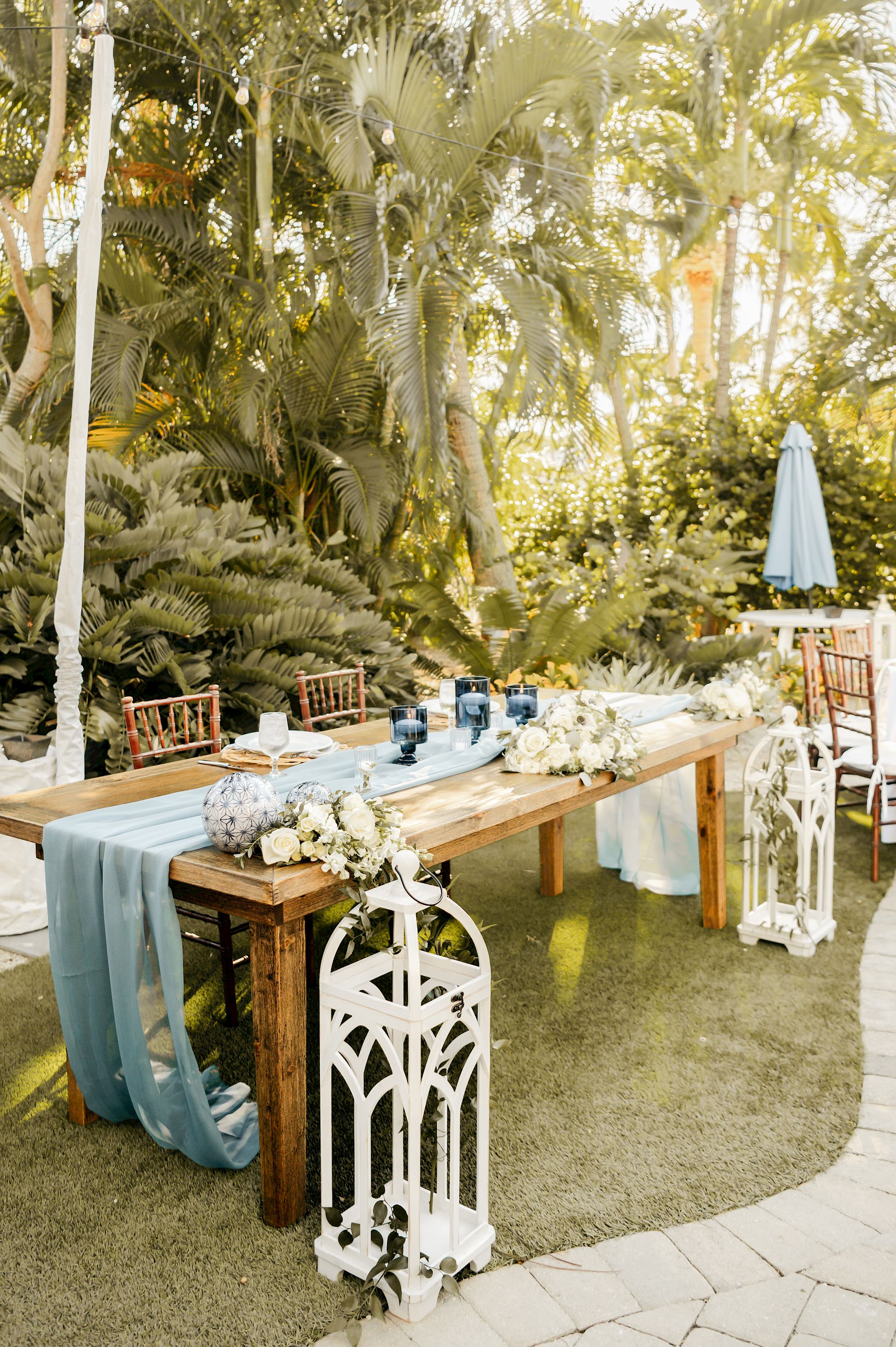 An outdoor table setup with blue decorations, flowers, and white lanterns, surrounded by lush greenery and string lights overhead.