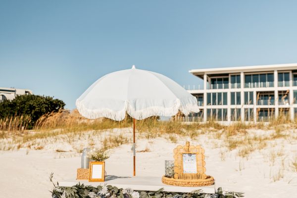 A decorated table with flowers and frames under a white parasol on a sunny beach, near a building.