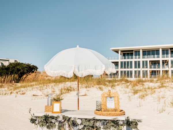 A decorated table with flowers and frames under a white parasol on a sunny beach, near a building.