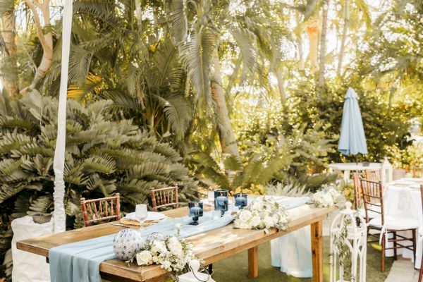 A rustic outdoor table setup with blue accents, white flowers, and lanterns in a tropical garden setting.